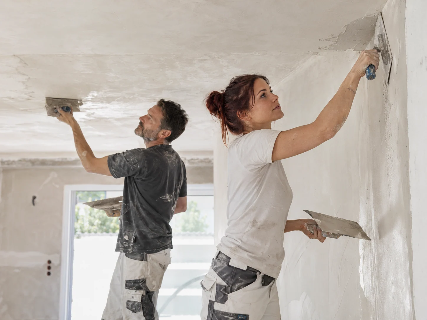 Stuckateurin und Kollege verputzen Wand und Decke eines Wohnraums